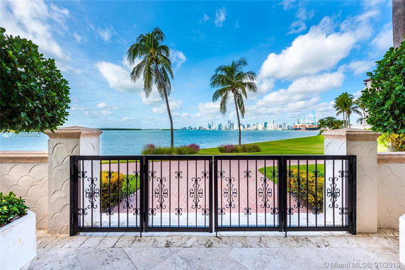 View through a decorative gate towards a waterfront with palm trees and a city skyline in the distance.