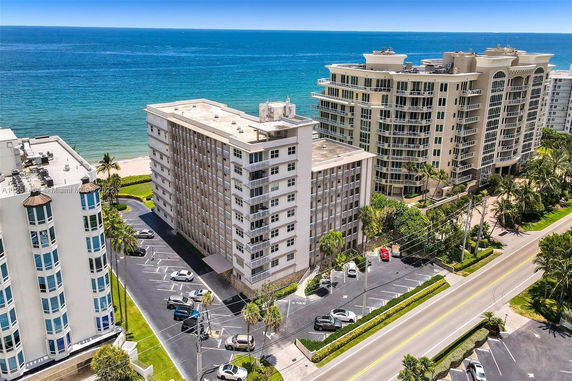 Aerial view of mid-rise beachfront condominium buildings with adjacent parking areas and the ocean in the background.