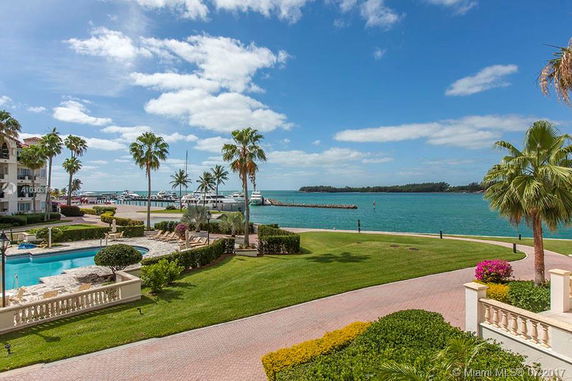 View of a waterfront area with a pool, landscaped gardens, palm trees, and a marina with boats.