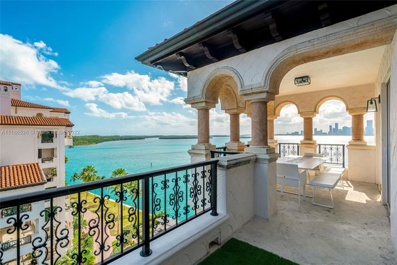 Balcony with arched columns and railing overlooking a body of water and city skyline.