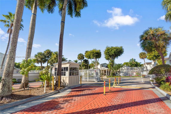 Entrance gate area with a security booth and road surrounded by palm trees.