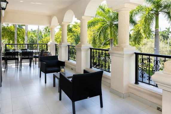 Covered outdoor terrace with black furniture and decorative columns, overlooking a garden with palm trees.