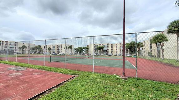 Tennis court surrounded by a chain-link fence with apartment buildings in the background.