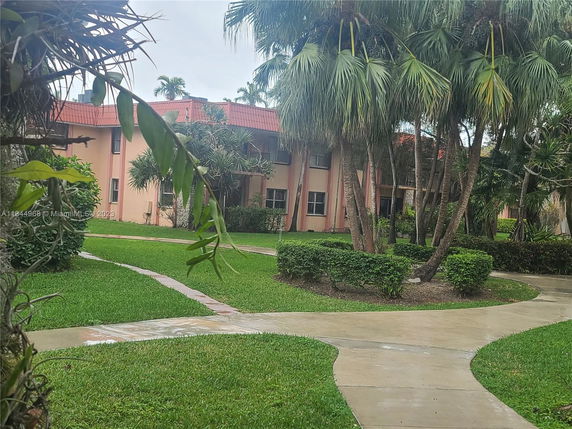 View of the front area of a house with a red-tiled roof, surrounded by a landscaped garden with palm trees and walkways.