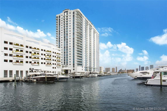 Front view of a high-rise residential building with multiple balconies, located near a water body with boats docked.