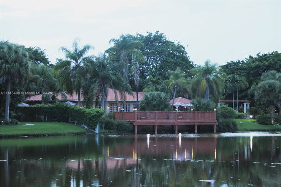 View of a house with red roof and wooden deck next to a body of water, surrounded by palm trees.