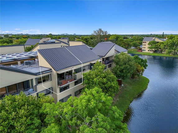 Aerial view of a multi-story residential building with balconies, situated near a body of water and surrounded by trees.
