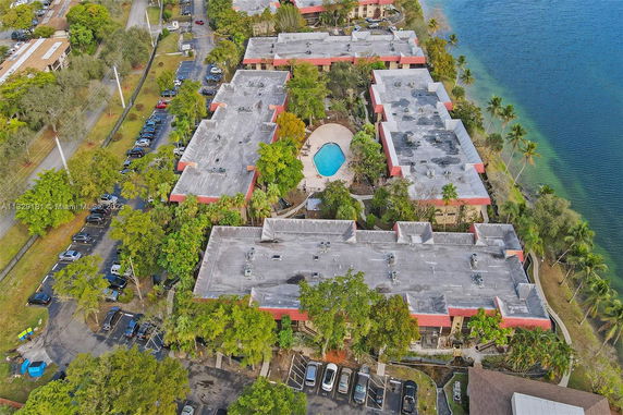 Aerial view of an apartment complex with multiple buildings, a swimming pool at the center, and parking areas, located next to a body of water.
