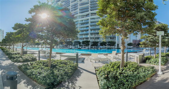 Outdoor swimming pool area with sun loungers and surrounding greenery in front of a multi-story building.