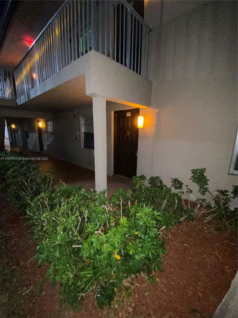 Night view of a building entrance with a balcony above and bushes in front.