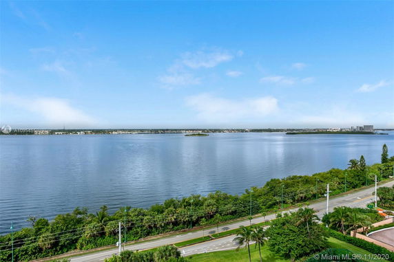 View of a water body with a road and greenery in the foreground, and buildings visible in the distance.