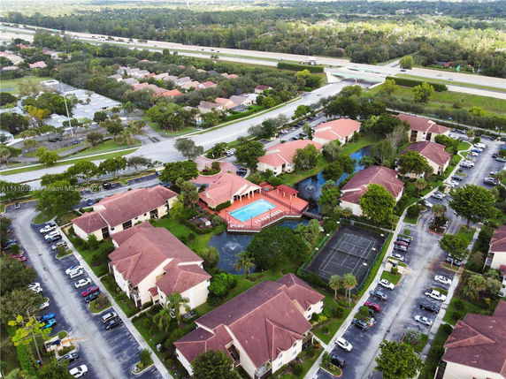 Aerial view of a residential complex with red-roofed buildings, a swimming pool, tennis court, and surrounding roads.