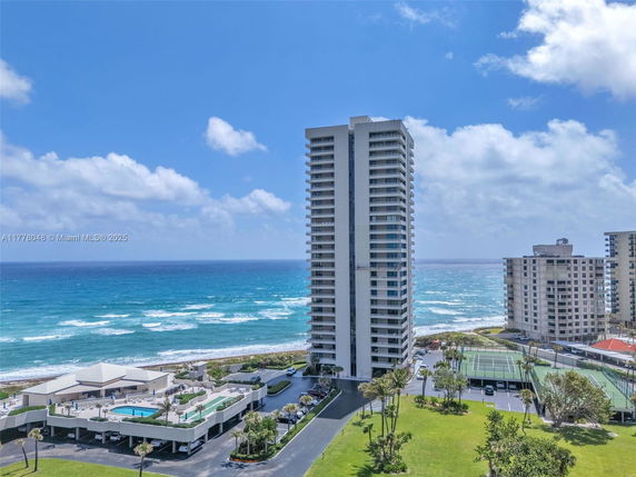 Front view of a tall residential building by the ocean with adjacent tennis courts and a swimming pool area on a bright day.