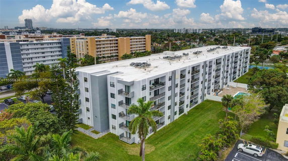 Aerial view of a long, multi-story residential building with balconies, surrounded by greenery and adjacent buildings.