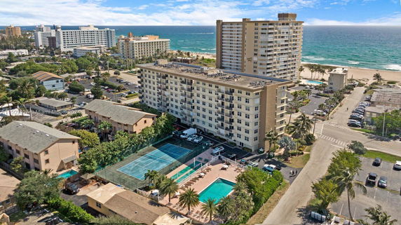 Aerial view of a coastal residential area with multiple buildings, including a high-rise and condominiums, with a tennis court and swimming pools, adjacent to a beach.