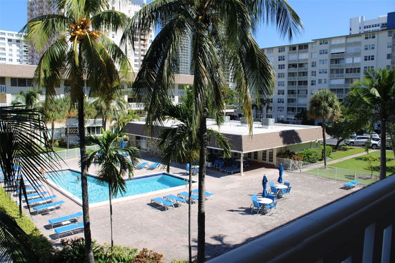 View of an outdoor swimming pool area surrounded by palm trees and adjacent buildings.