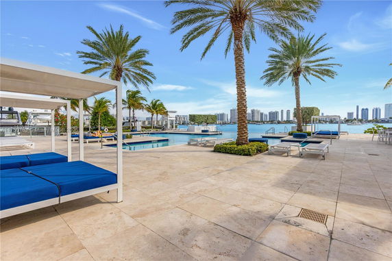 Outdoor swimming pool area with lounge chairs and palm trees, overlooking a body of water and distant city skyline.