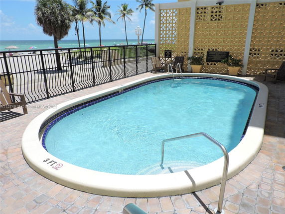 Outdoor swimming pool near a beach with a decorative wall and palm trees in the background.