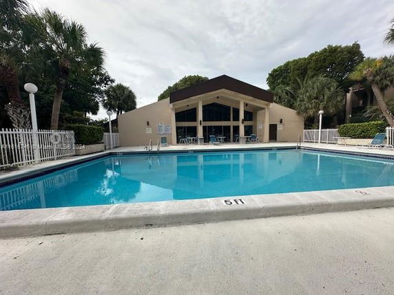 Outdoor swimming pool in front of a building with a fenced perimeter and surrounding trees.