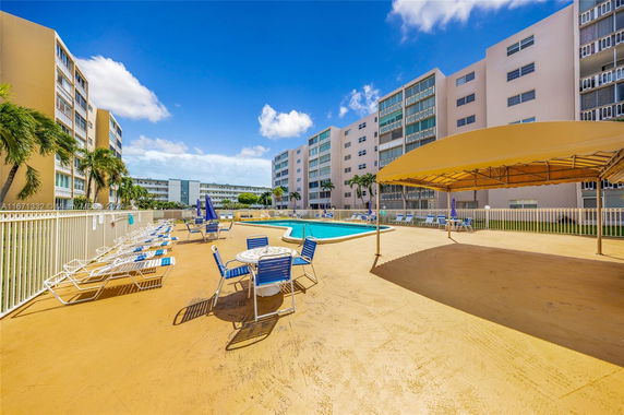 Outdoor swimming pool area with lounge chairs and umbrellas surrounded by multi-story buildings.