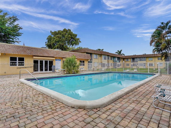 Outdoor swimming pool with surrounding deck area and two-story building in the background.