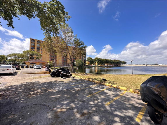 View of a multi-story building near a water body, with parked vehicles and a tree-lined area.
