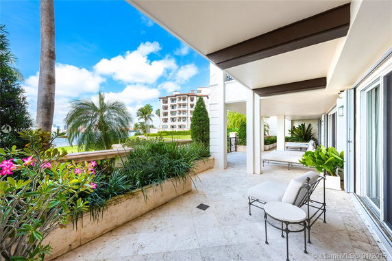 Outdoor view from a covered patio with seating, overlooking a garden area with palm trees and a multi-story building in the background.