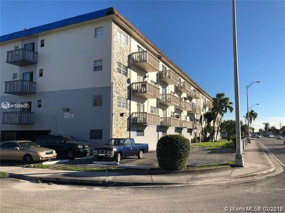 Corner view of a multi-story apartment building with balconies and a parking area in front.