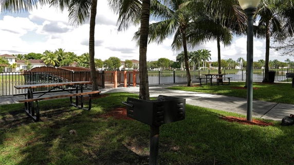 Park area with picnic tables, a barbecue grill, and views of a pond, enclosed by palm trees and a fence.