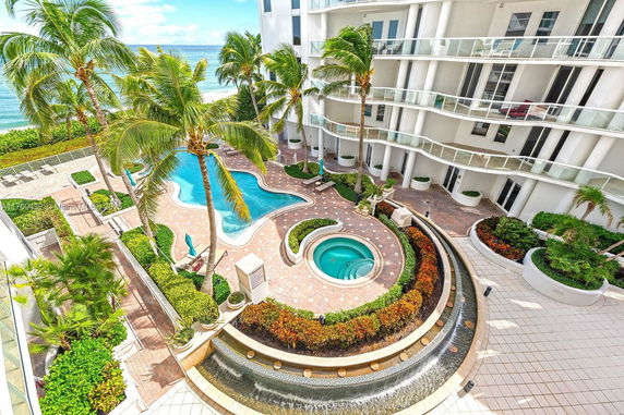 Outdoor swimming pool area with palm trees and surrounding tall building.
