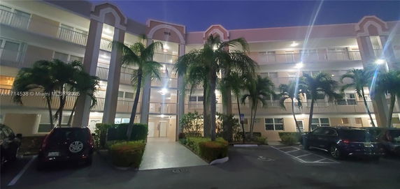 Evening view of a multi-story building with balconies and a nearby parking lot with several cars.