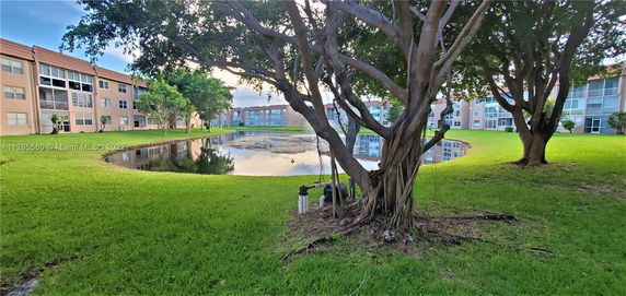 View of a residential building complex with a pond and trees in the foreground.