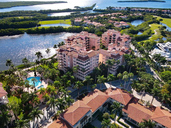Panoramic aerial view of a waterfront residential complex with pink buildings and a swimming pool surrounded by palm trees.