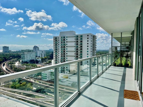 Balcony view of high-rise buildings with a glass railing and a wide cityscape visible under a blue sky with clouds.