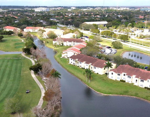 Aerial view of residential buildings near a river with green lawns and surrounding landscape.