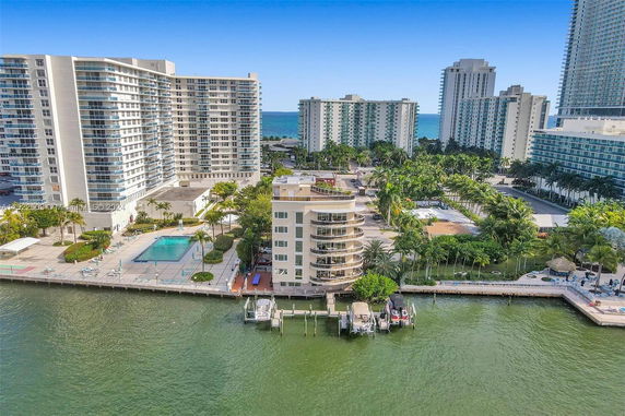 Aerial view of a waterfront building complex with a swimming pool, surrounded by water and high-rise buildings.