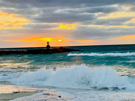 Beach view at sunset with waves and a distant lighthouse on a rocky jetty.