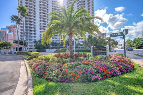 Front view of a tall residential building with balconies, surrounded by landscaped gardens and a palm tree.