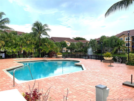 Outdoor swimming pool area with surrounding palm trees and multi-story buildings in the background.
