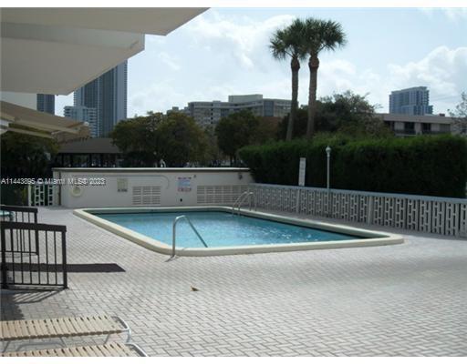 Outdoor swimming pool area with a nearby hedge and trees, surrounded by high-rise buildings in the background.