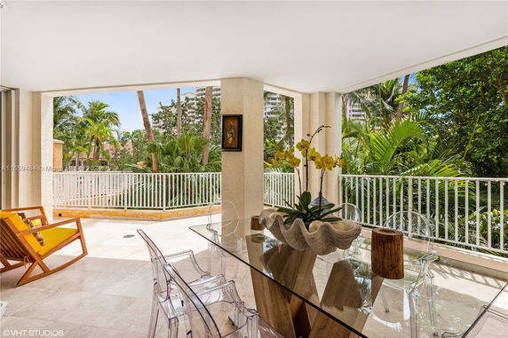 Outdoor patio area with glass table and chairs, surrounded by greenery and visible buildings in the background.