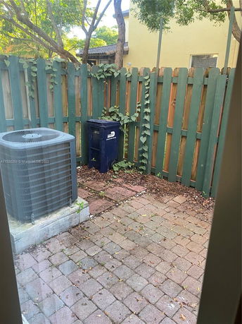 Outdoor area with a small paved section, surrounded by a wooden fence, an air conditioning unit, and a trash bin.