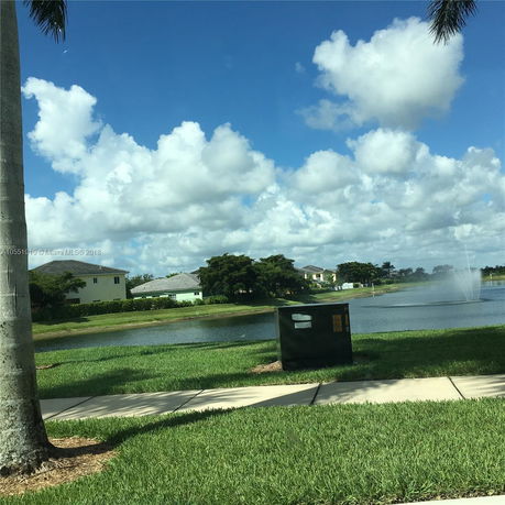 View of a pond with houses and a fountain in the distance, under a cloudy sky.