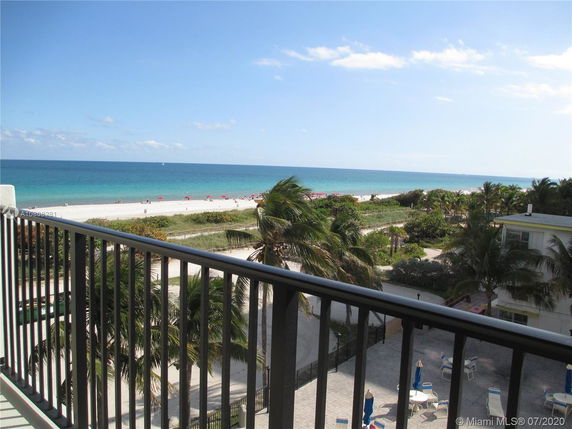 View of the ocean and beach from a balcony, with palm trees and a building visible below.