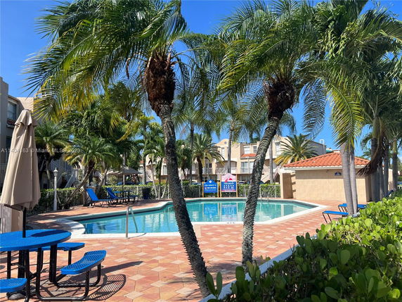 Outdoor swimming pool area with adjacent tables and lounge chairs, surrounded by palm trees.