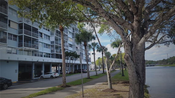 Side view of a multi-story building with balconies, adjacent to a sidewalk and a river.