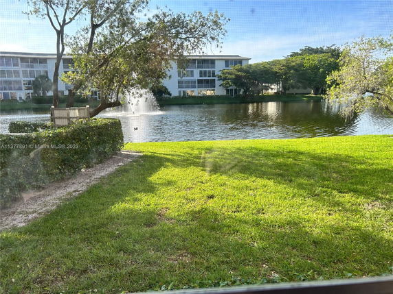 View of a pond with a fountain and surrounding trees, with a multi-story building in the background.