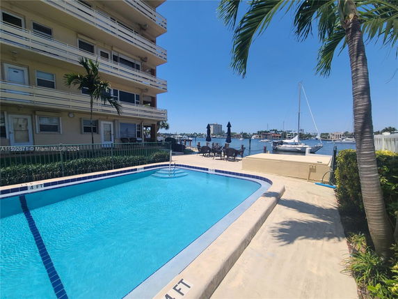 View of a multi-story building with balconies adjacent to a swimming pool and waterfront area with boats.