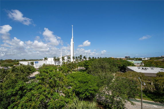 Wide view of a white building with a tall spire surrounded by trees and palm trees.
