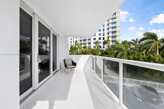 Balcony with glass railing and outdoor seating, overlooking nearby trees and a modern high-rise building.